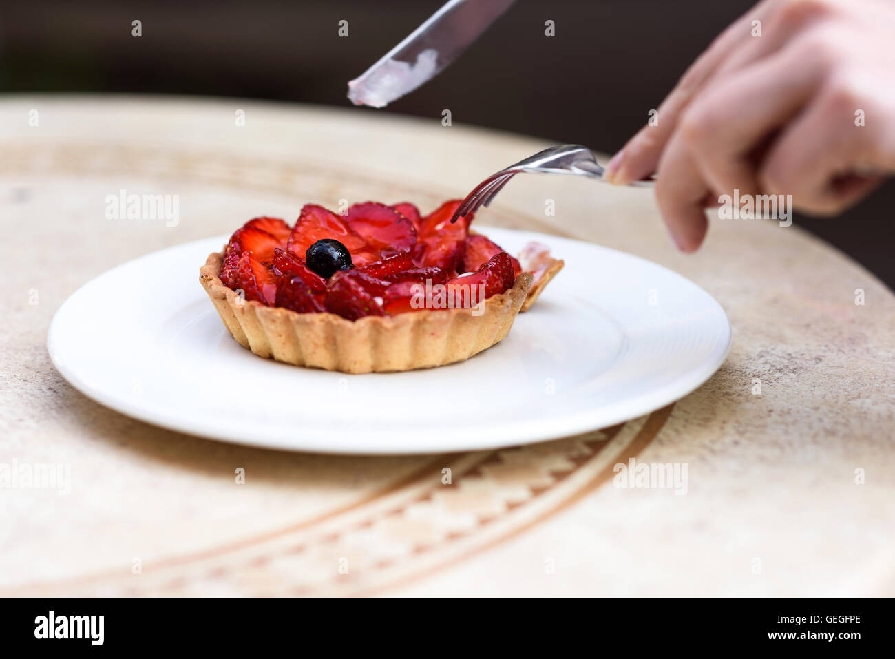 Photo of female hands cutting a fruit cake with strawberries, mint and blackcurrant, on a white plate on a restaurant table. Stock Photo