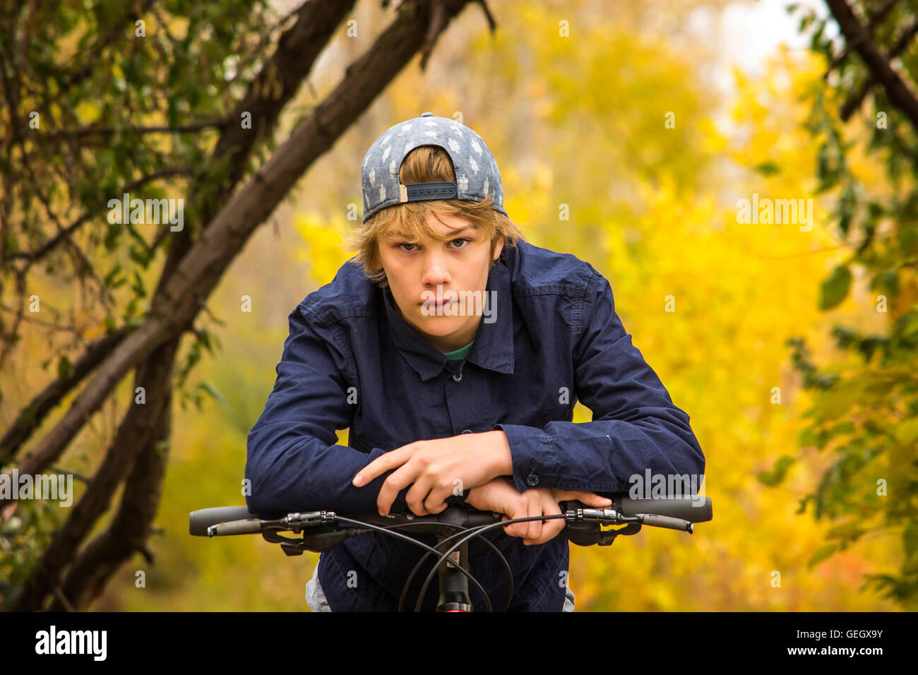 a teenage boy is resting his arms on the handle bar of his bicycle, posing for the camera in front of a brigt colored trees back Stock Photo