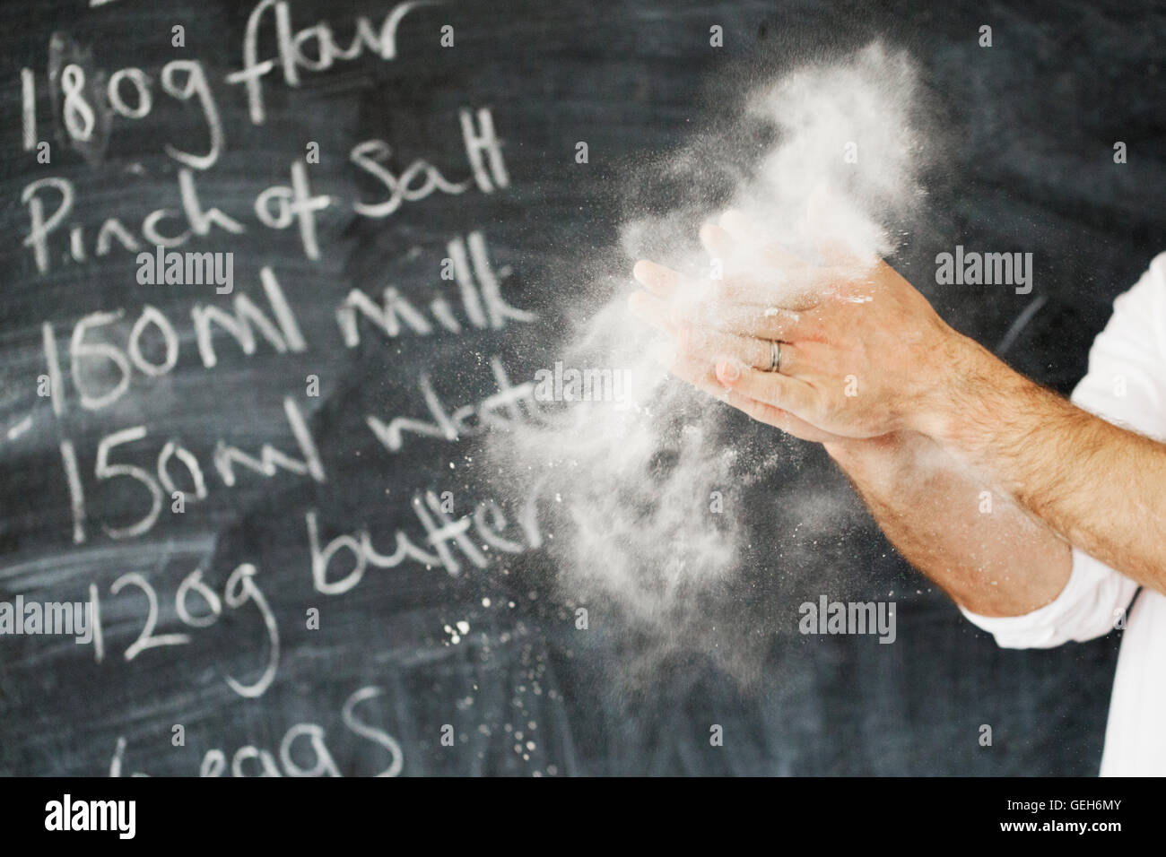 Close up of a baker standing in front of a blackboard, dusting his hands with flour. Stock Photo
