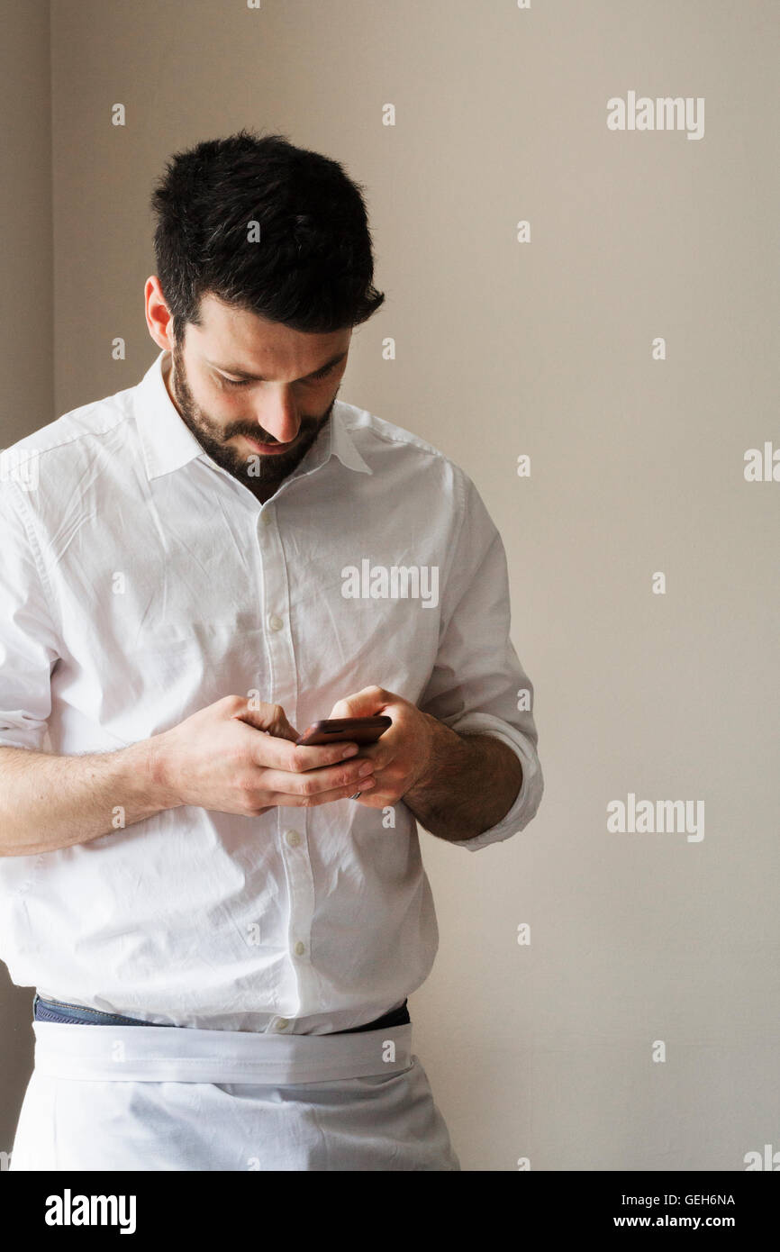 Half length portrait of a bearded man wearing a white apron, using his smart phone. Stock Photo