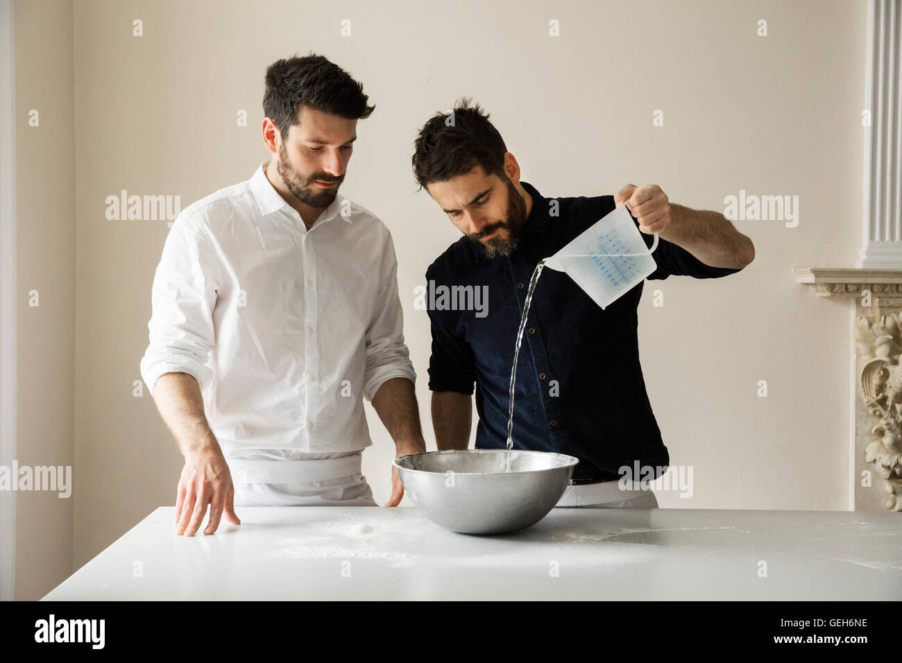 Two bakers standing at a table, preparing bread dough, pouring water from a measuring jug into a metal mixing bowl. Stock Photo