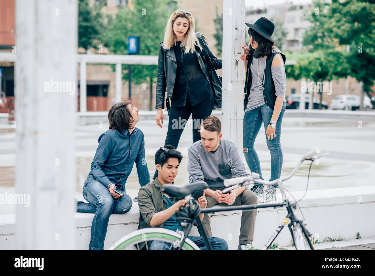 Group of young multiethnic friends sitting on a small wall talking to each other, using a smartphone, having fun - friendship, relaxing concept Stock Photo