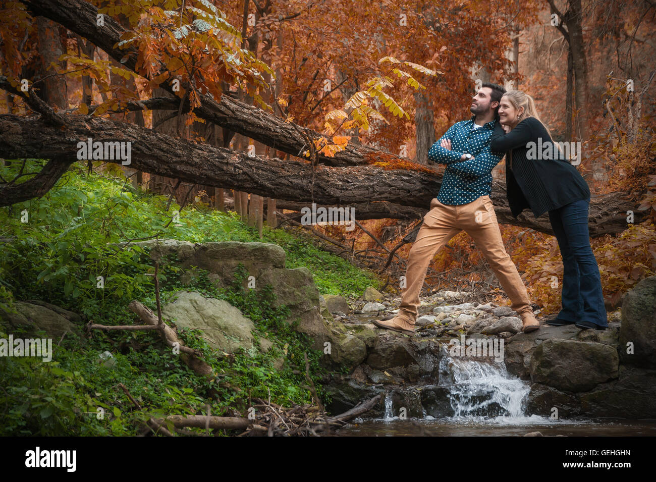 Couple cuddle next to a large tree branch and a stream in autumn Stock Photo