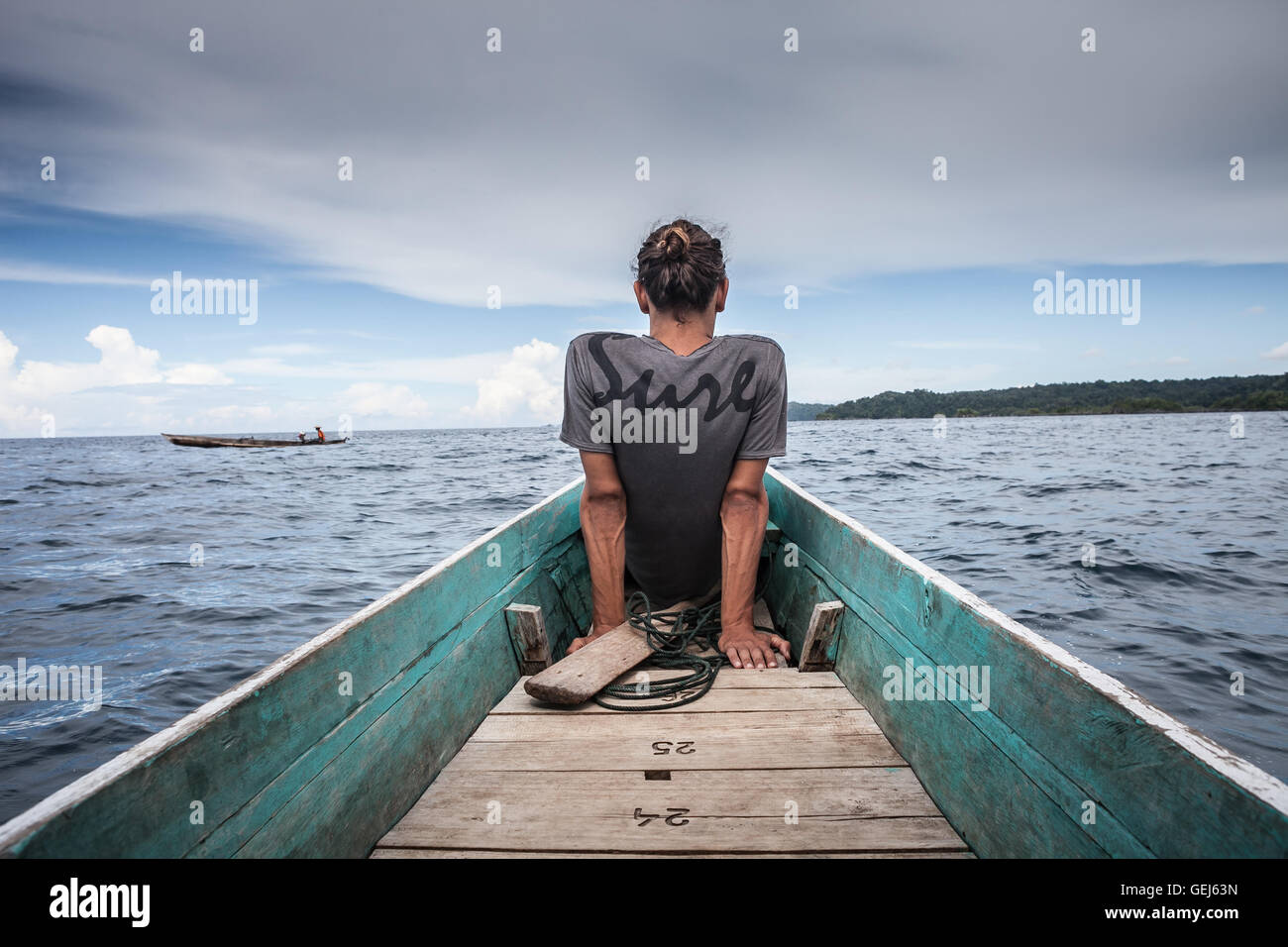 Photo of Man Sitting Natural Wood Long Tail Boat Caribbean Ocean. Clear water and blue sky with clouds. Horizontal. Stock Photo