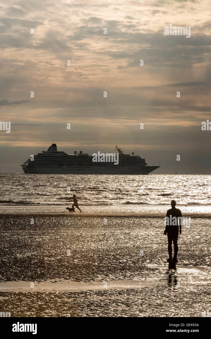 Crosby, Liverpool, Merseyside, UK. 27th July, 2016. People running on the beach in the evening sun, beside one of the Antony Gormley Iron Men statues that make up the  'Another Place' installation, as the Crystal Cruise Line ship Crystal Symphony leaves Liverpool after it's first visit to Liverpool thisa year. Crystal cruise CEO Edie Rodriguez was heard on Radio Four Today programme on Wednesday Ju;y 27th talking to John Humphrys about  their plans to run cruises through the North West Passage in August 2016. Credit:  John Davidson Photos/Alamy Live News Stock Photo