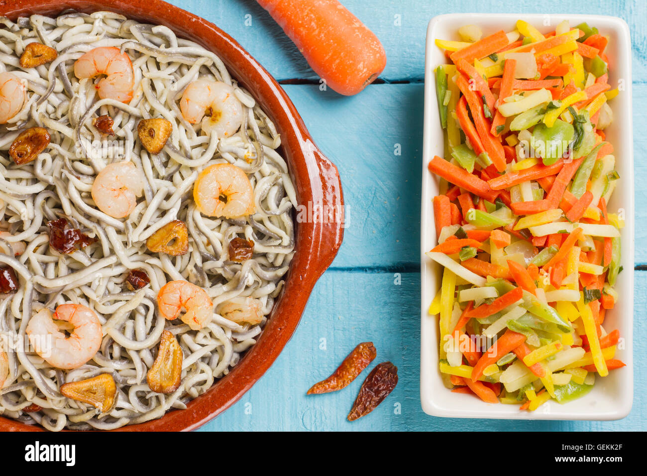 Surimi elvers with prawns, garlic and pepper on a clay pot, vegetables on a white bowl, carrot and peppers on a wooden table Stock Photo