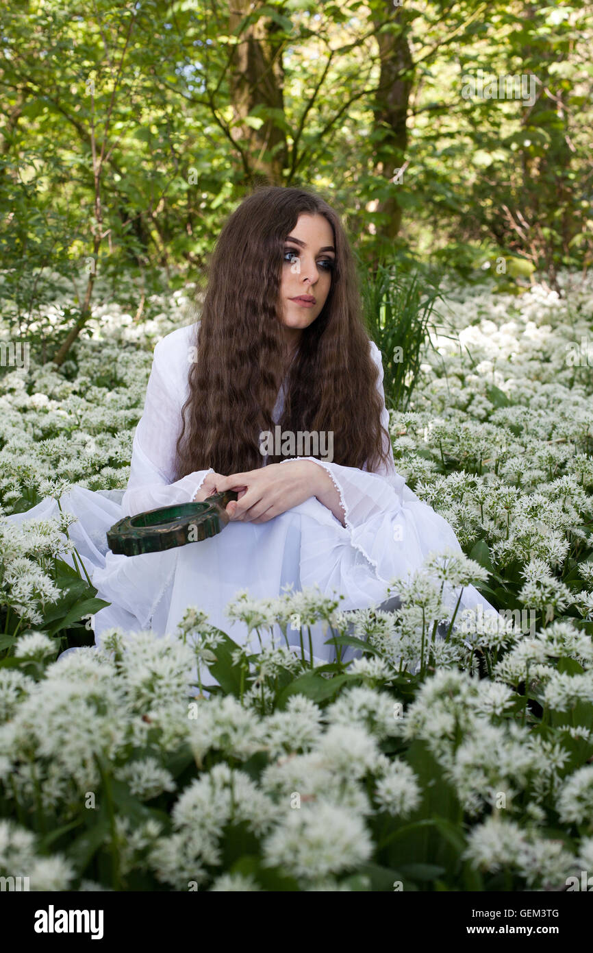 Beautiful woman wearing a long white dress sitting amongst white flowers holding a mirror Stock Photo