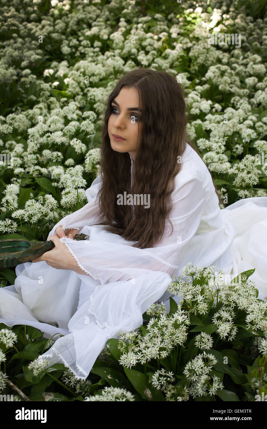 Beautiful woman wearing a long white dress sitting amongst white flowers holding a mirror Stock Photo