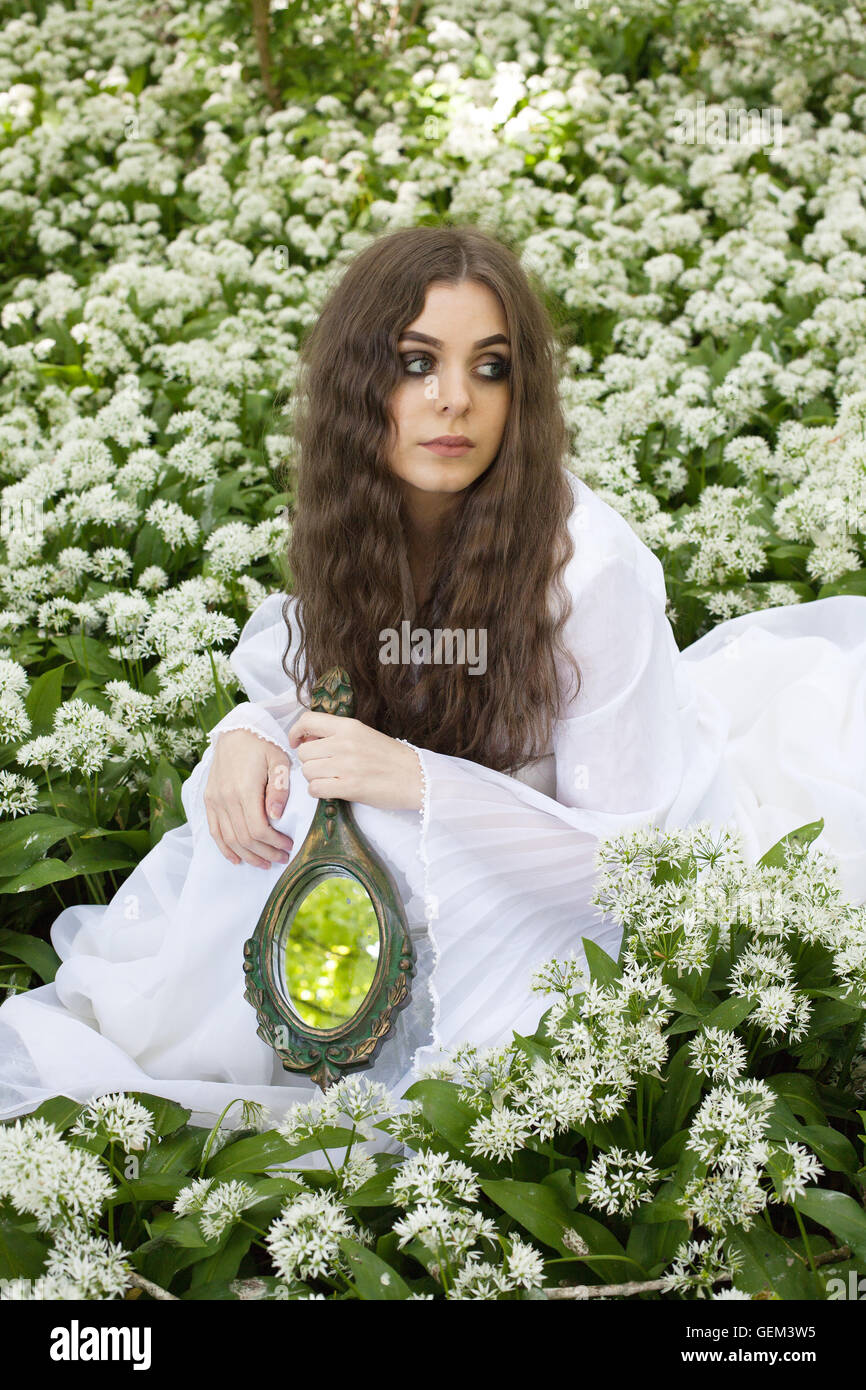 Beautiful woman wearing a long white dress sitting amongst white flowers holding a mirror Stock Photo