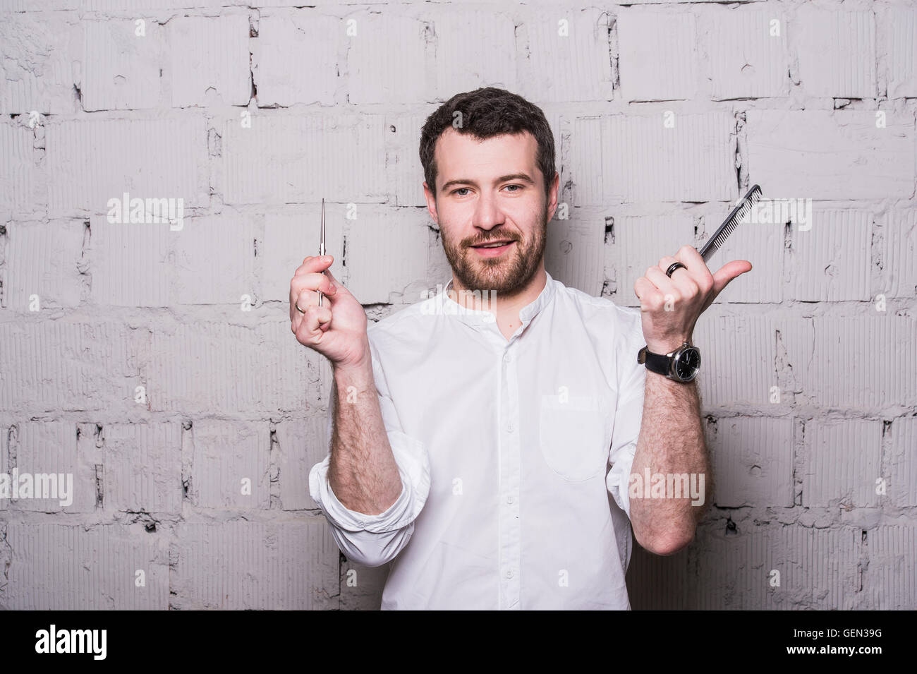 Young hairdresser, wearing in white shirt, posing with scissors and comb, on grey brick wall background Stock Photo