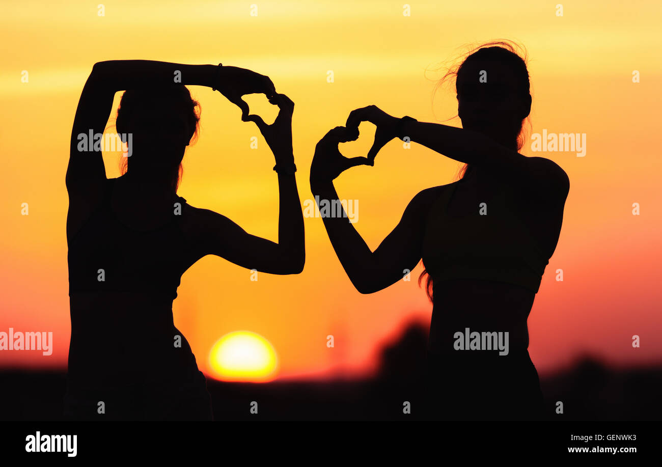 Landscape with silhouette of young sporty women holding hands in heart shape on the background of colorful sky at sunset Stock Photo