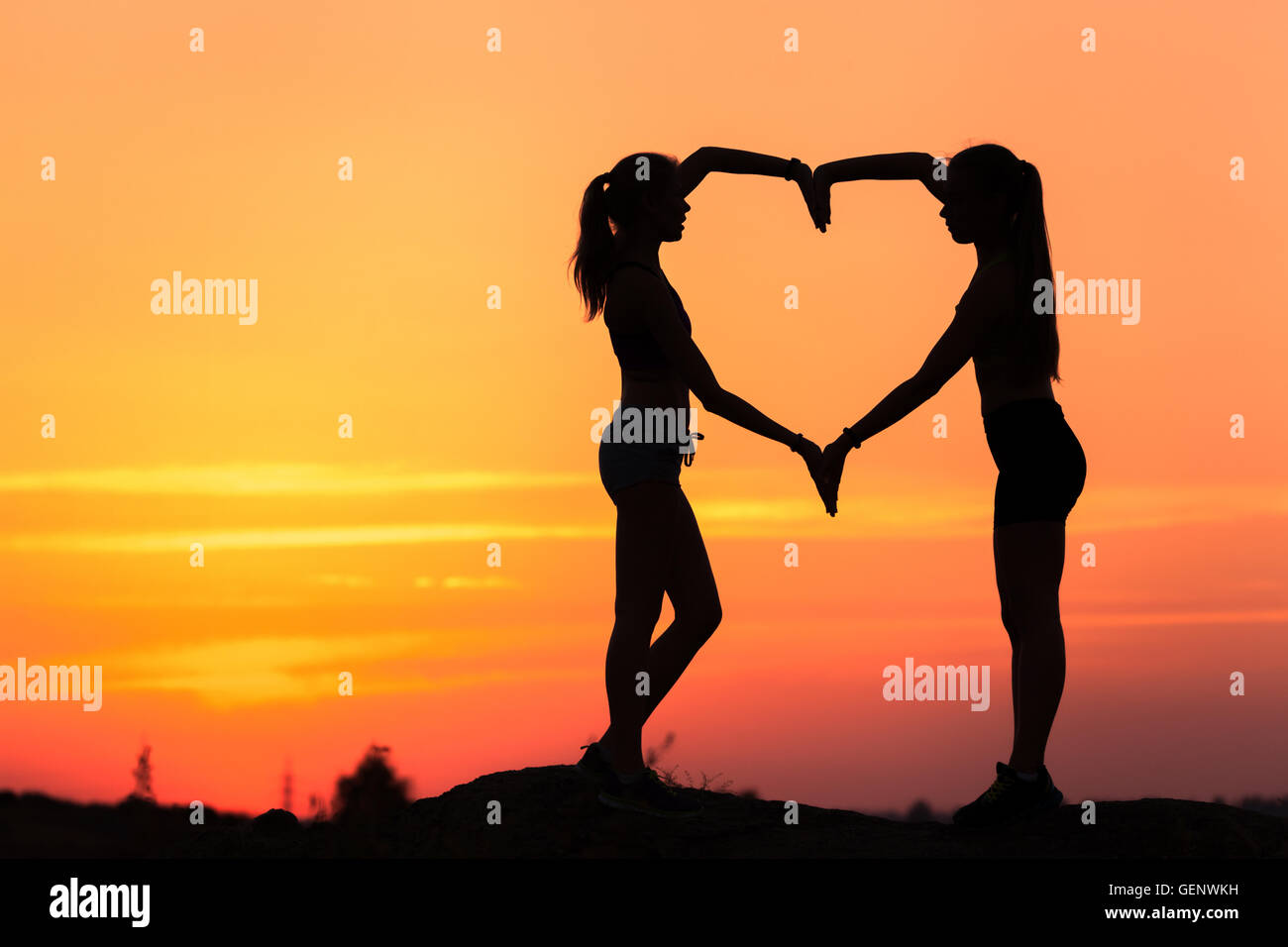 Landscape with silhouette of young sporty women holding hands in heart shape on the background of colorful sky at sunset in summ Stock Photo