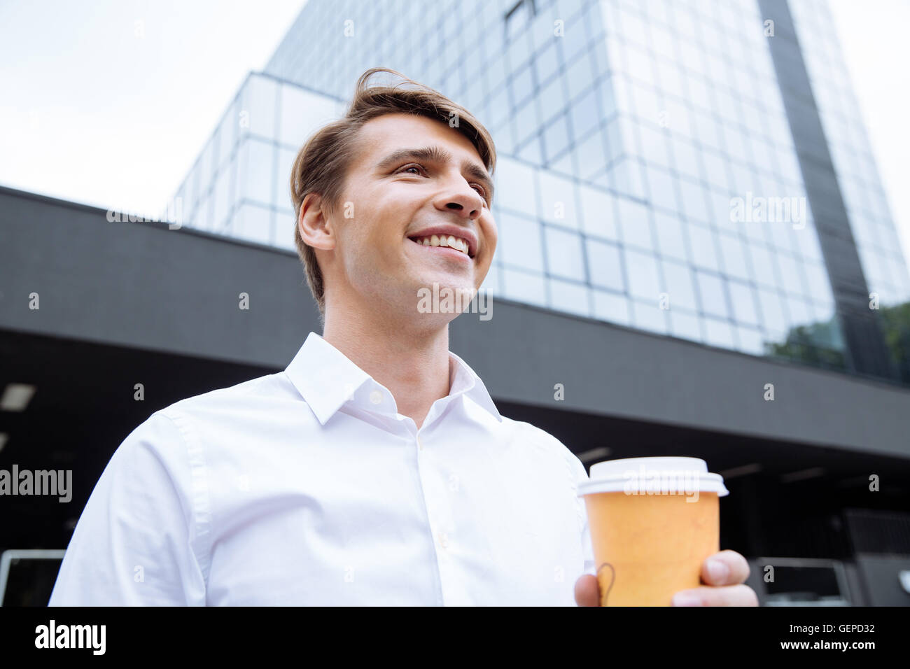Happy confident young businessman standing near skyscraper and drinking ...