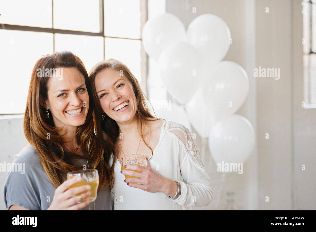 Two women standing side by side holding drinks. White balloons, party decorations. Stock Photo