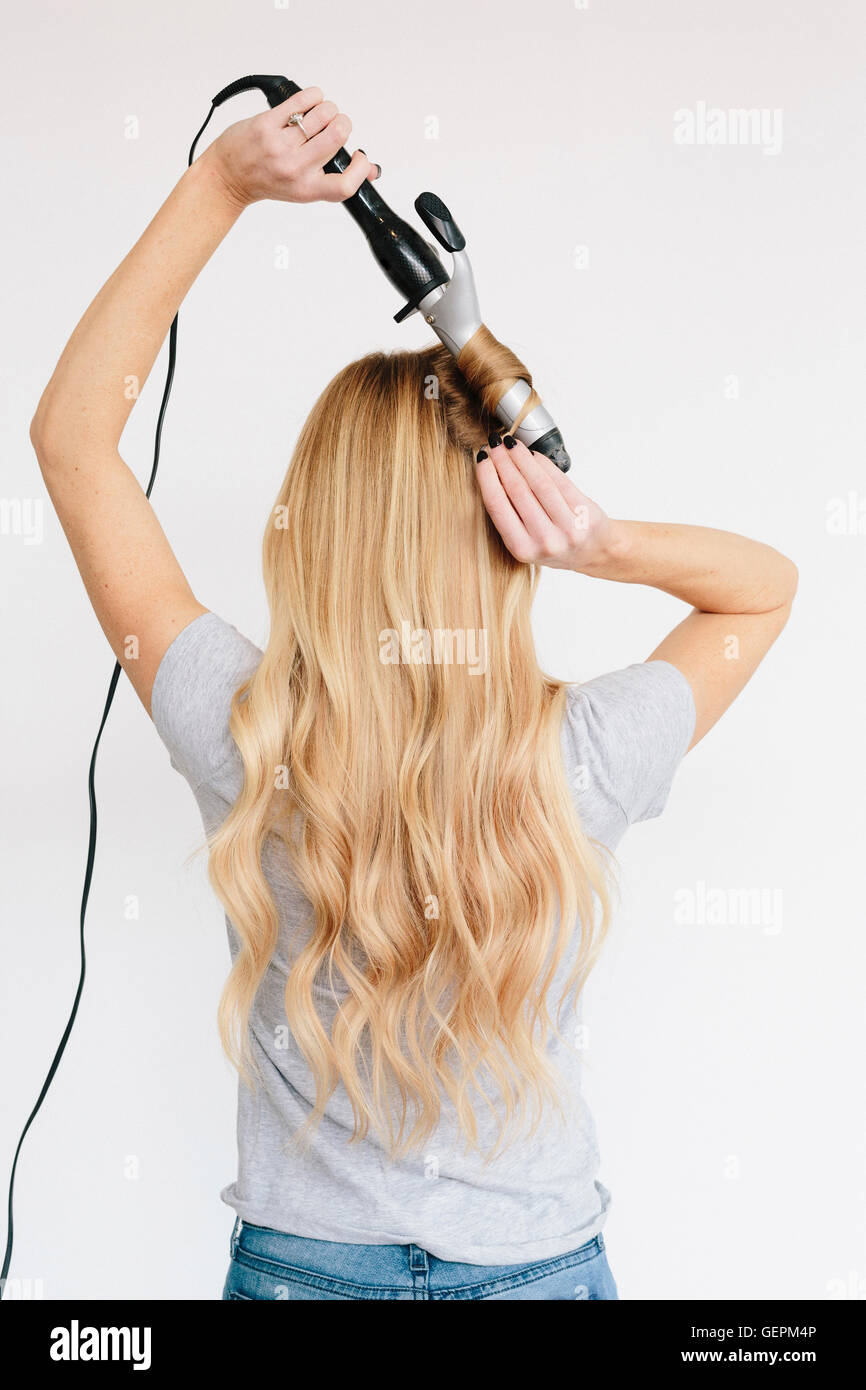 A young woman with long blond wavy hair using curling tongs. Back view. Stock Photo