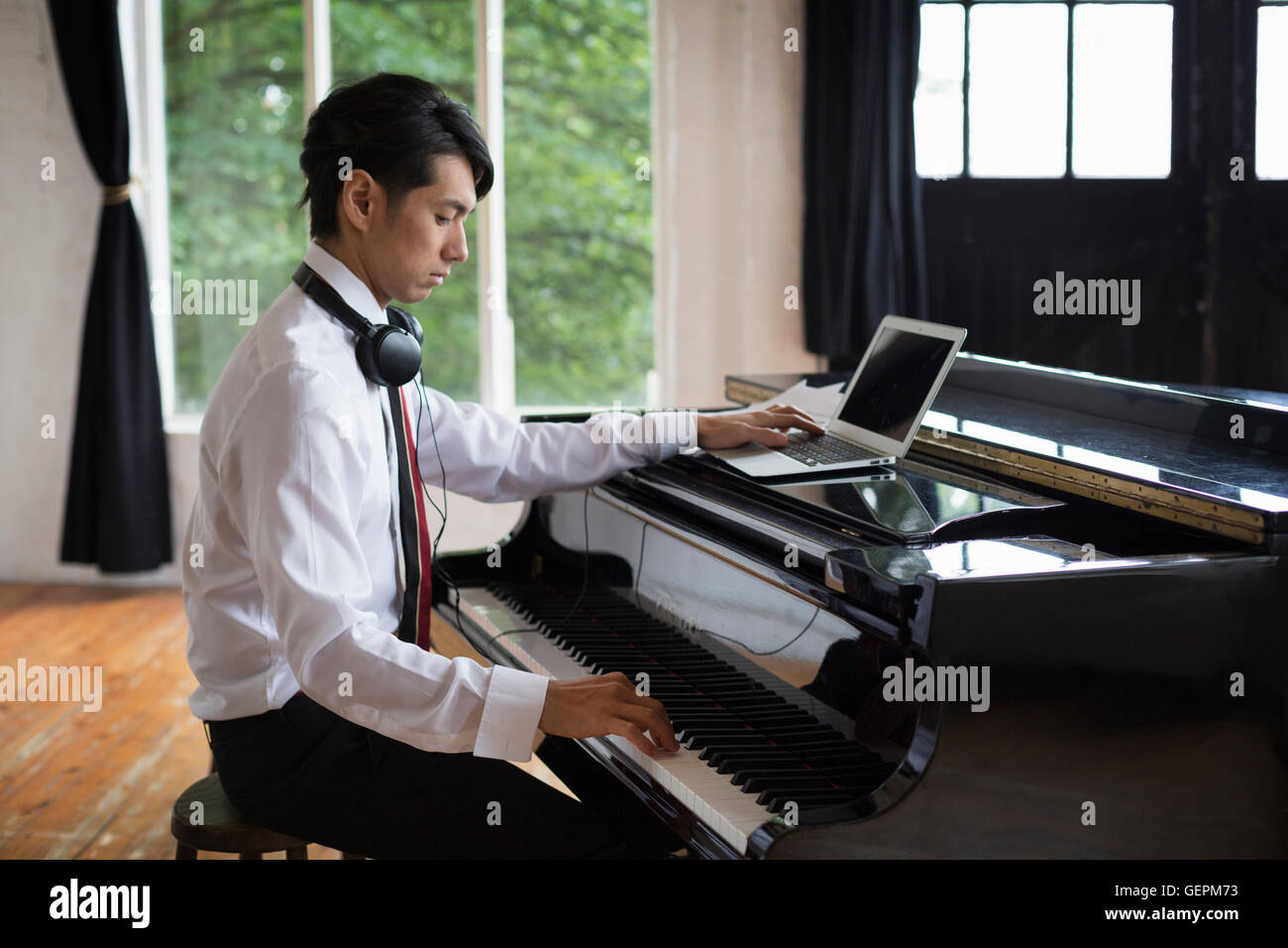 Young man sitting at a grand piano in a rehearsal studio, playing and simultaneously using a laptop computer. Stock Photo