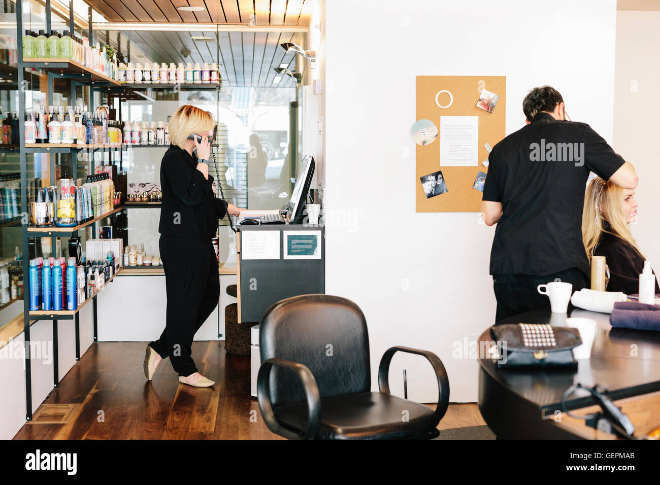 A hair stylist working on a client's hair, and a woman on the telephone, looking at a laptop computer. Stock Photo