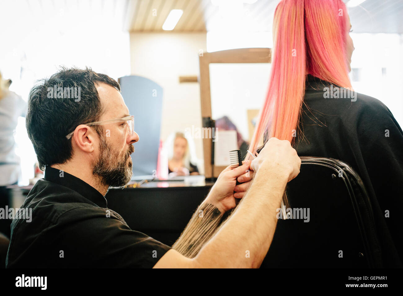 A hair stylist with a client, trimming the ends of her long dyed pink straight hair. Stock Photo