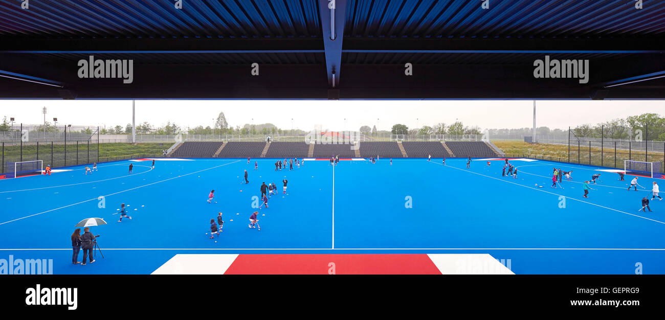 Hockey field in blue with canopied spectator stand. Eton Manor - Lee Valley Hockey and Tennis Centre, London, United Kingdom. Architect: Stanton Williams, 2014. Stock Photo
