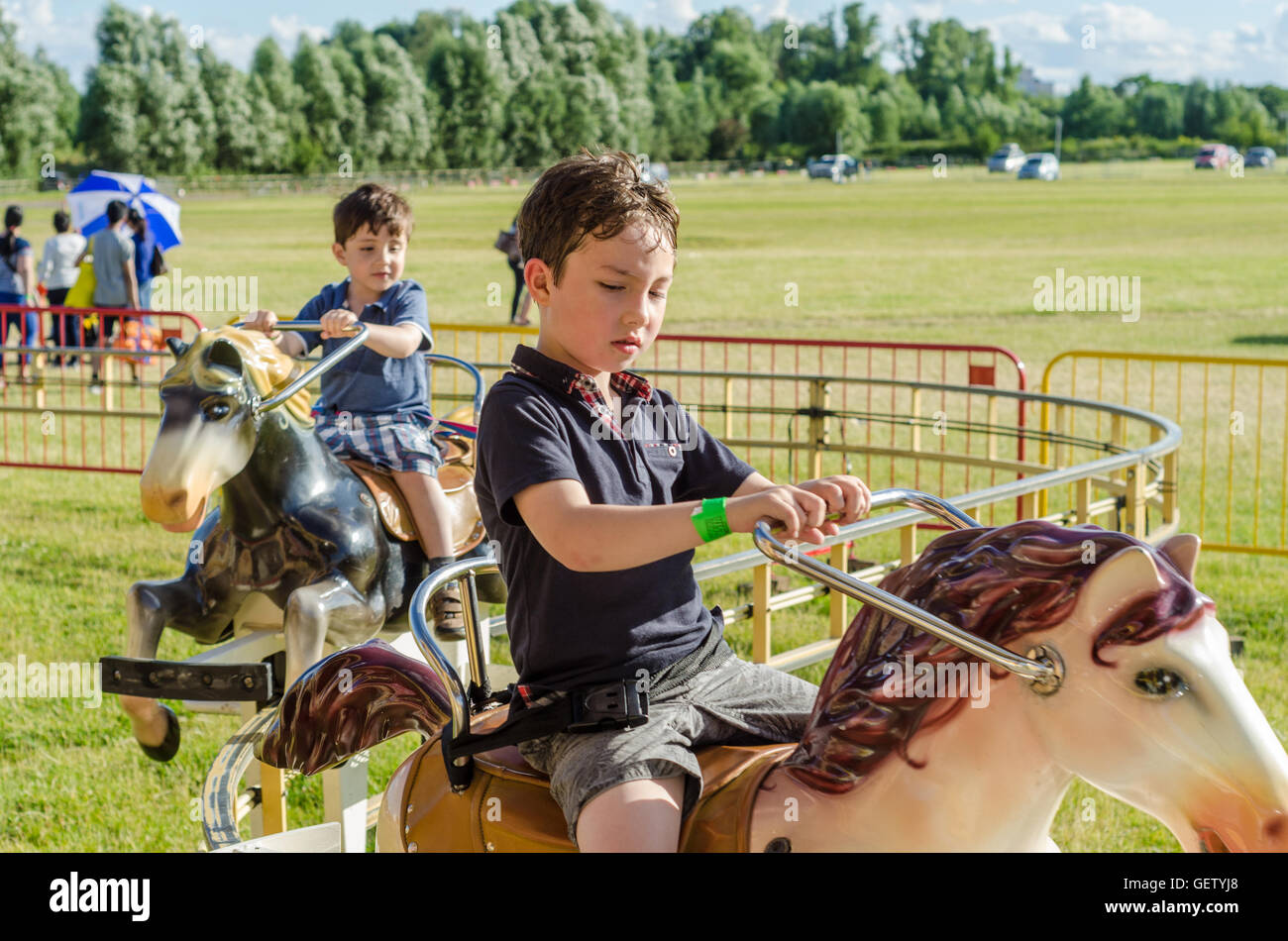 Two young boys ride on a ride at a fairground where they sit on toy horses and ride around a track. Stock Photo