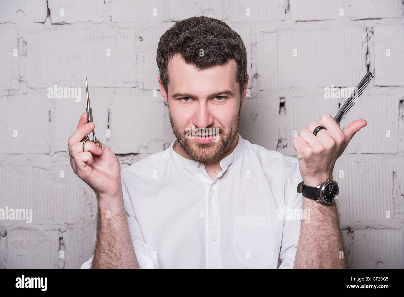Young hairdresser, wearing in white shirt, posing with scissors and comb, on grey brick wall background Stock Photo