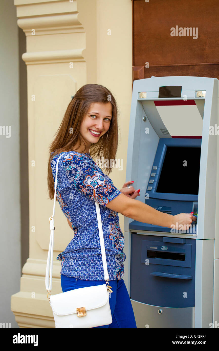 Smiling young woman in a blue blouse withdrawing money from ATM Stock Photo