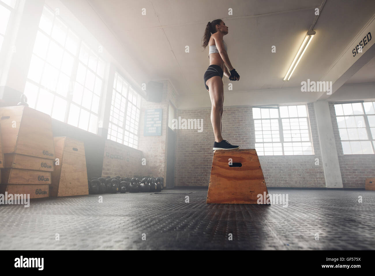 Low angle shot of fit young female model in sports wear standing on a box at gym. Muscular woman doing box jump exercise at gym. Stock Photo