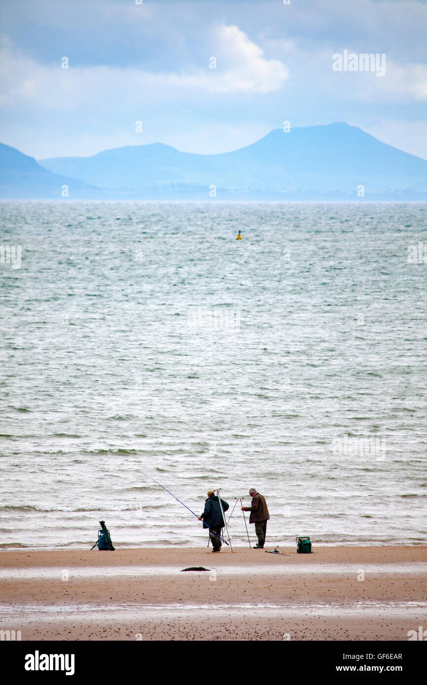 Two fishermen fishing on the beach at Newborough Warren, Anglesey, Wales, UK with the Llyn Peninsula coastal mountains in the distance Stock Photo