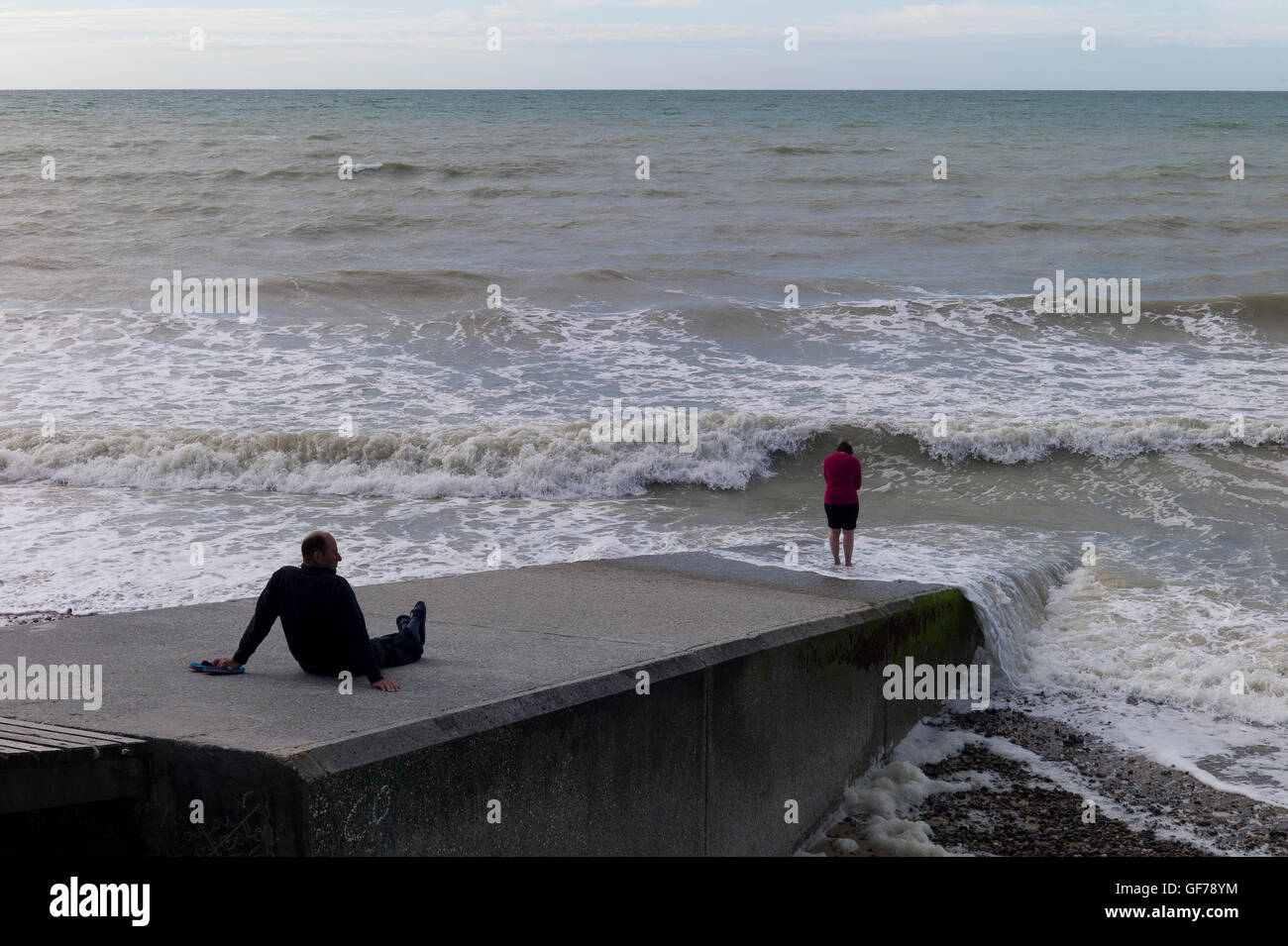 Man sitting on a breakwater watching his wife paddle in the sea, Normandy,France Stock Photo