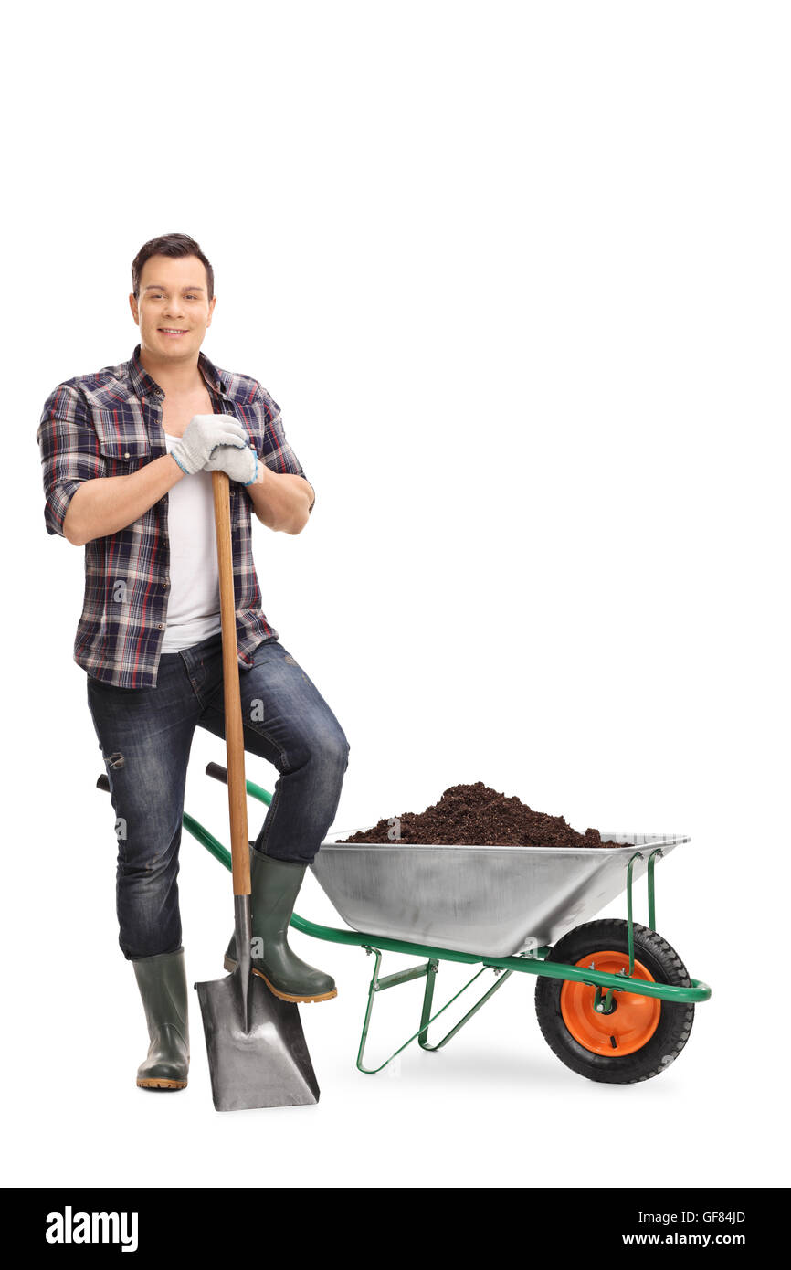 Full length portrait of a young gardener posing with a shovel by a wheelbarrow with dirt isolated on white background Stock Photo