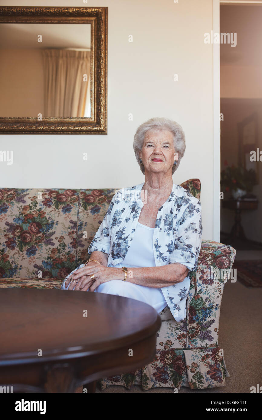 Portrait of an elderly woman sitting on a couch at old age home. Caucasian woman sitting on a sofa and looking at camera. Stock Photo