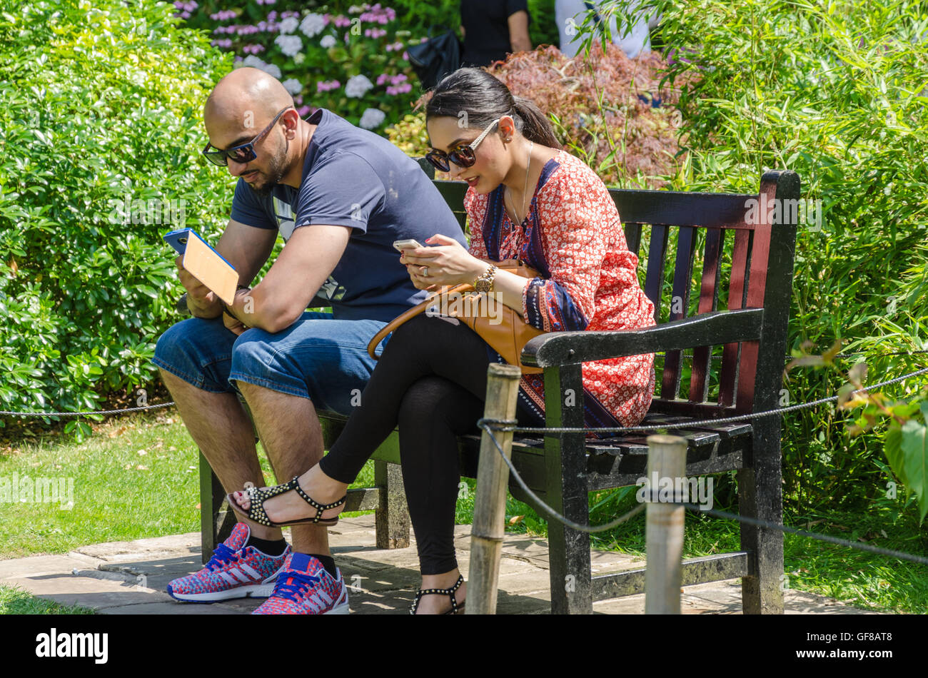 A couple sit together on a bench in Holland Park, London and use their smartphones. Stock Photo