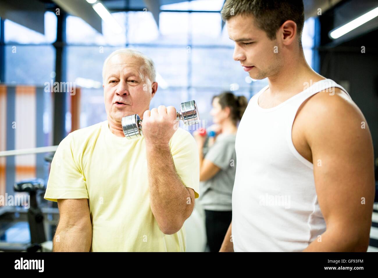 PROPERTY RELEASED. MODEL RELEASED. Senior man picking up dumbbells with trainer standing in gym. Stock Photo