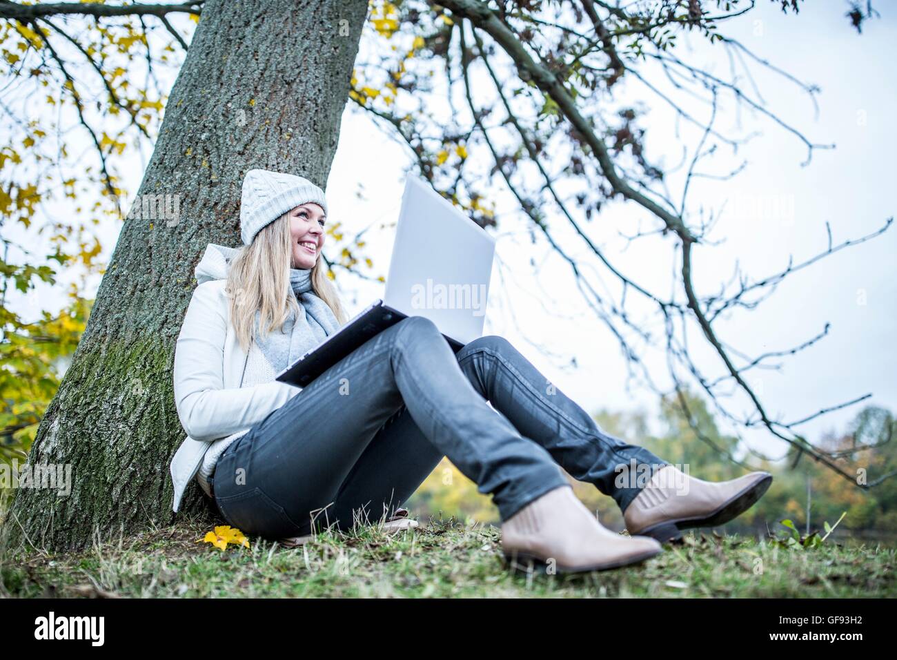 MODEL RELEASED. Young woman sitting at tree trunk and using laptop. Stock Photo