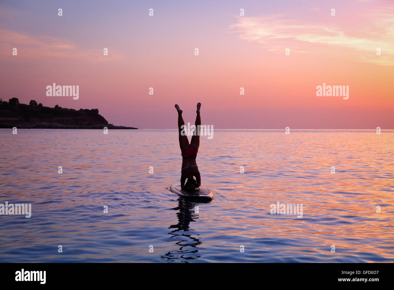 Silhouette of a sportive woman doing yoga exercise on the sup board over sunset, doing Sirsasana, headstand, active lifestyle Stock Photo