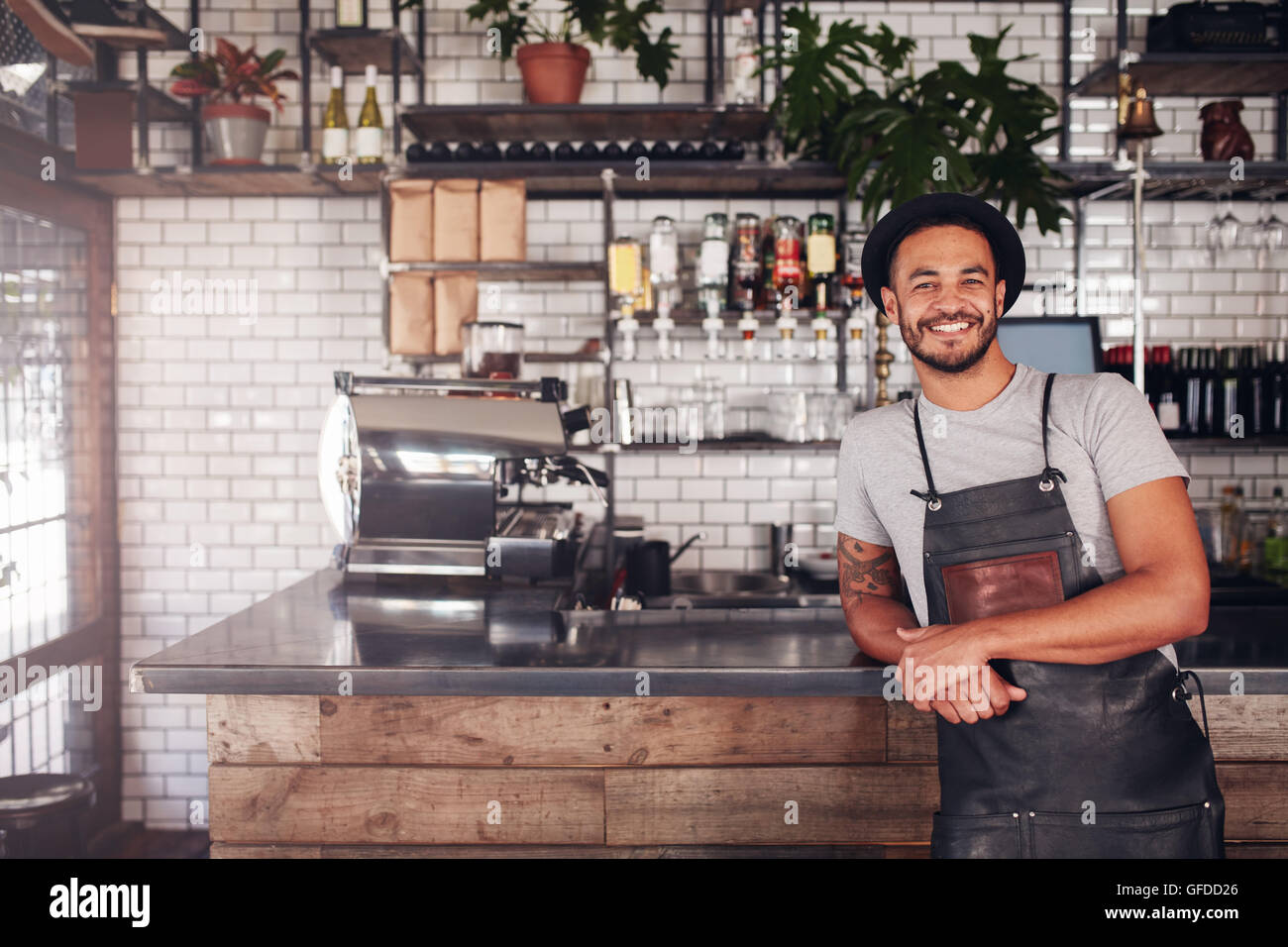Portrait of young man standing at the counter in his cafe. Coffee shop working in apron and hat smiling at camera. Stock Photo
