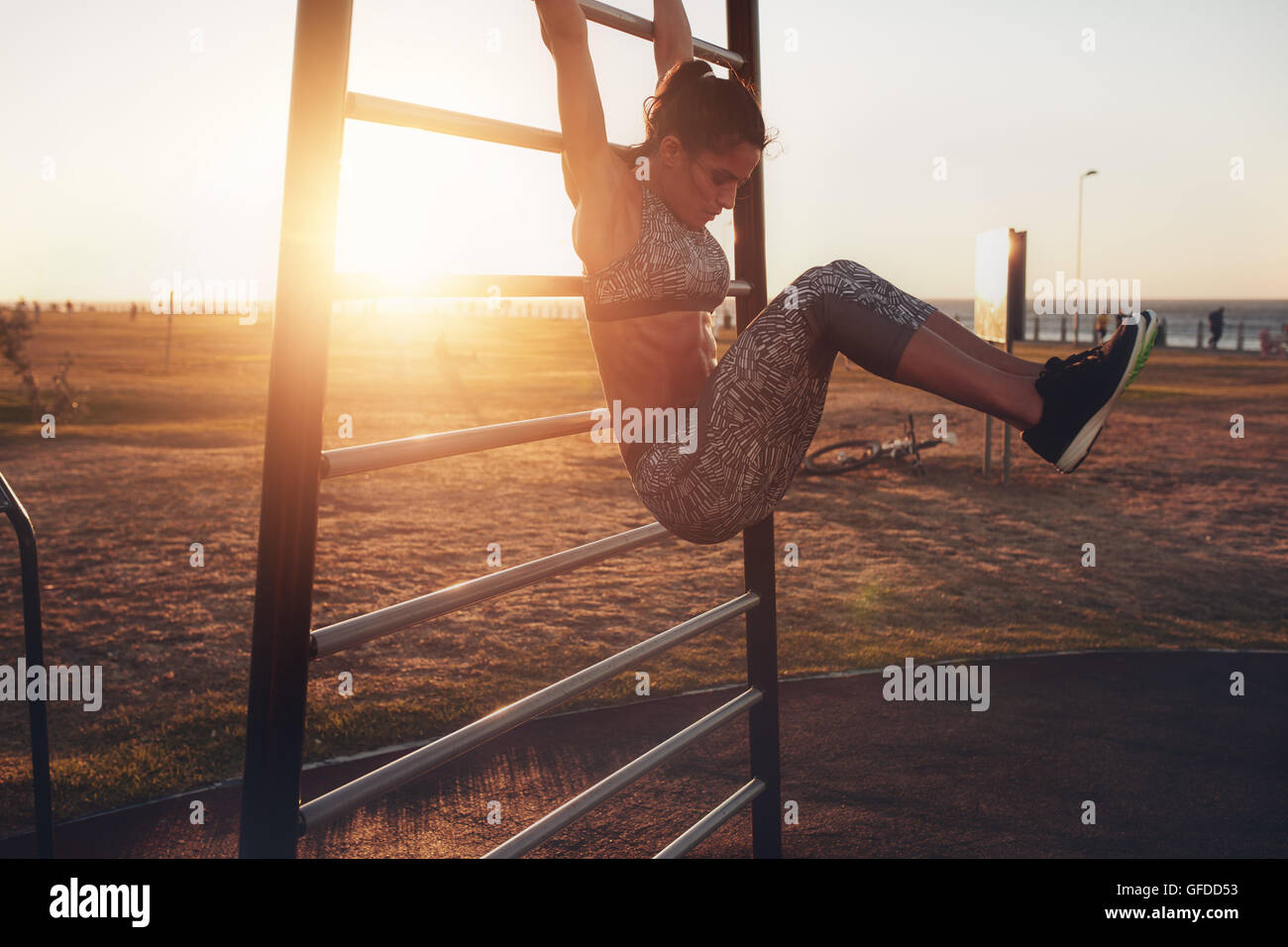 Candid shot of real healthy and fit woman performing hanging leg raises on outdoor fitness station in sunset at beach promenade. Stock Photo