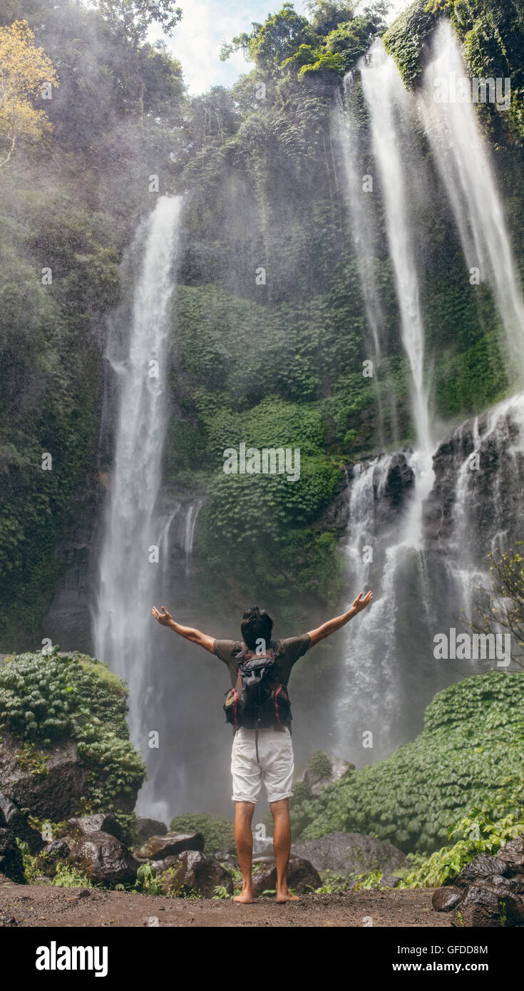 Rear view of young man standing in front of waterfall with his hands raised. Male tourist with arms outstretched enjoying near w Stock Photo
