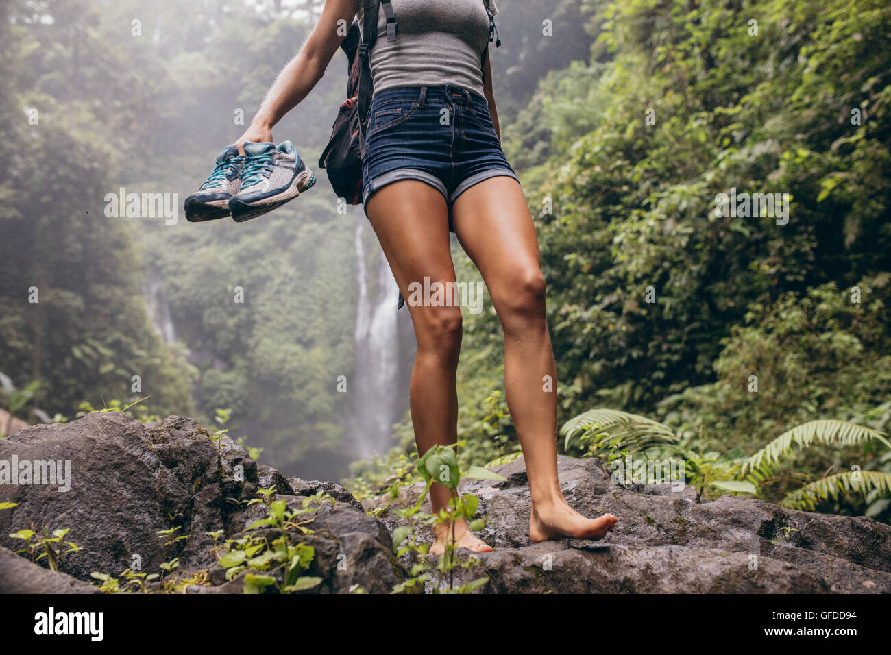 Low angle view of young woman walking down the mountain barefoot. Female hiker walking barefoot on rock with waterfall in backgr Stock Photo