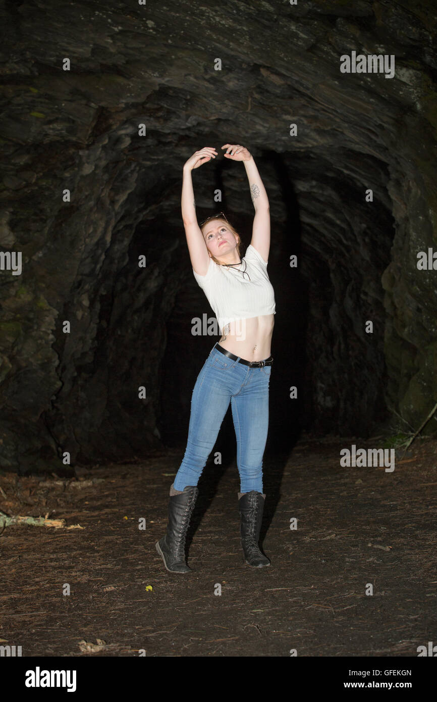 Beautiful red head in skinny jeans, black boots and white top, standing with arms raised at the mouth of a granite tunnel. Stock Photo