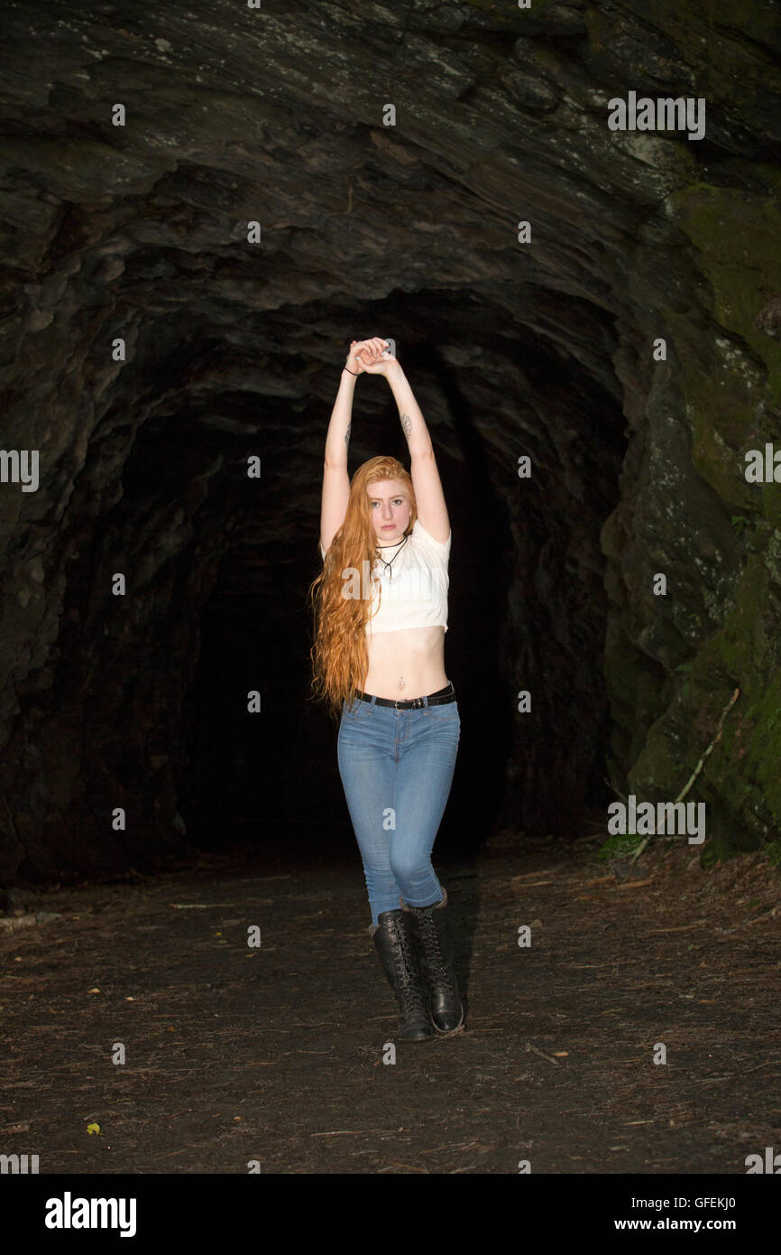 Beautiful red head in skinny jeans, black boots and white top, standing with arms raised at the mouth of a granite tunnel. Stock Photo
