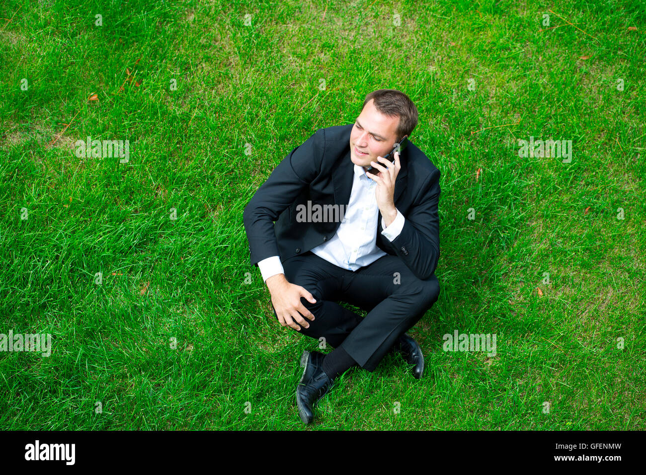 Cheerful young businessman calling by phone sitting on green grass Stock Photo