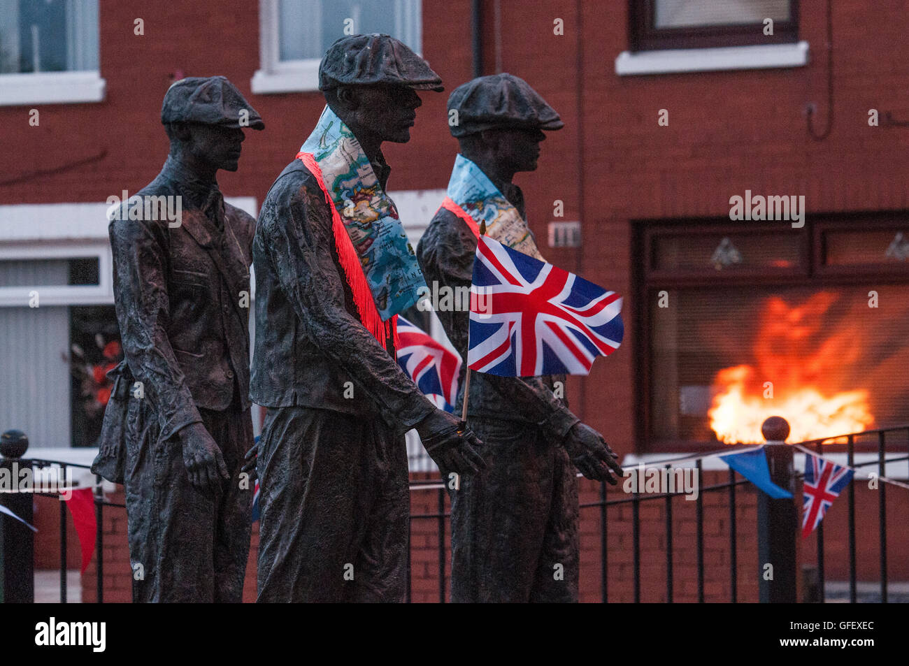Belfast, Northern Ireland. 11 Jul 2016 - The reflection of a bonfire in a window behind a public art display remembering the shipyard workers of Belfast, which has been decorated with Orange 'Sashes' or collarettes and Union Flags in readiness for the 12th July celebrations Stock Photo