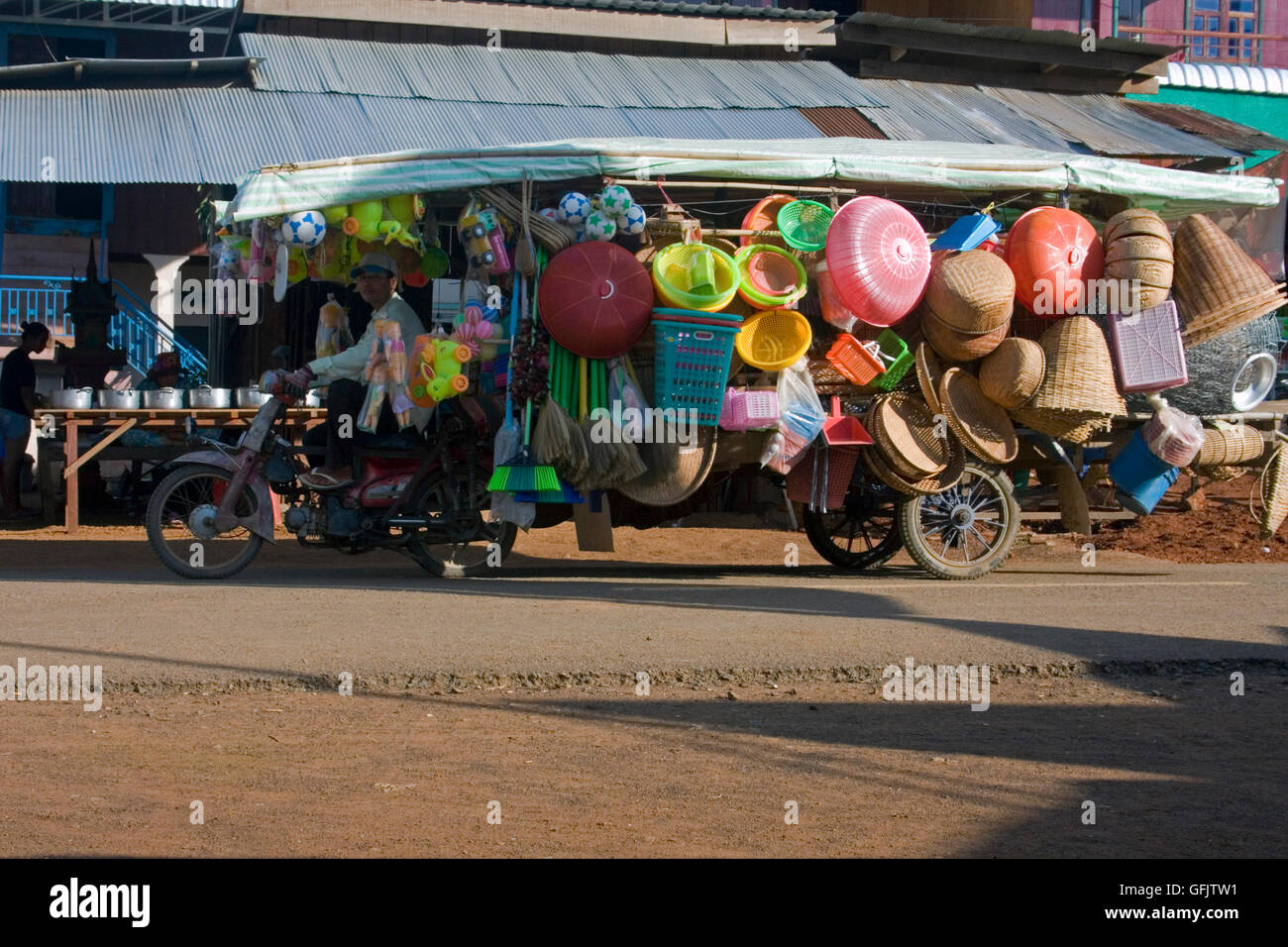 A man is driving a motorcycle towing a trailer loaded with toys & household items on a city street in Chork village, Cambodia. Stock Photo