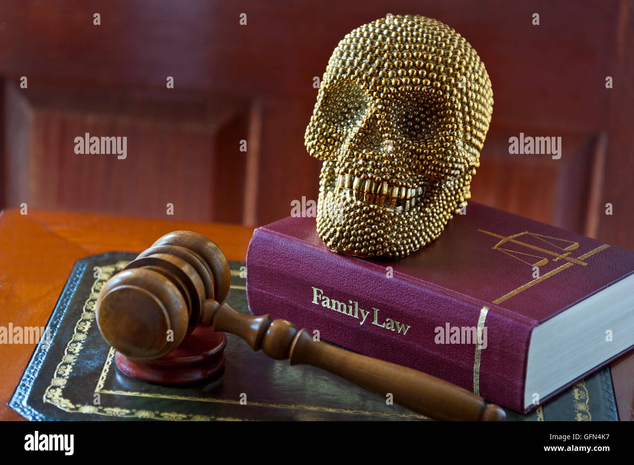 Smiling gold themed human like skull on Family Law book with judges gavel on inlaid desk ‘The last laugh’ Stock Photo