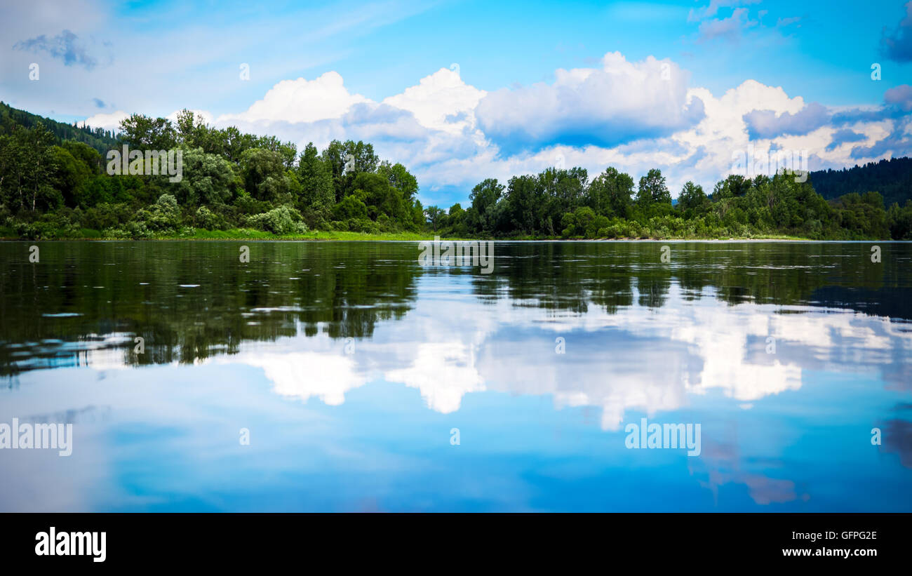 Beautiful landscape panorama with cloudy blue sky reflected in the clear water. Wooded waterside of a mountain lake. Stock Photo