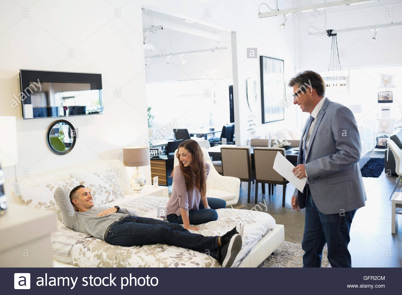 Salesman helping couple laying on bed in home furnishings store Stock
