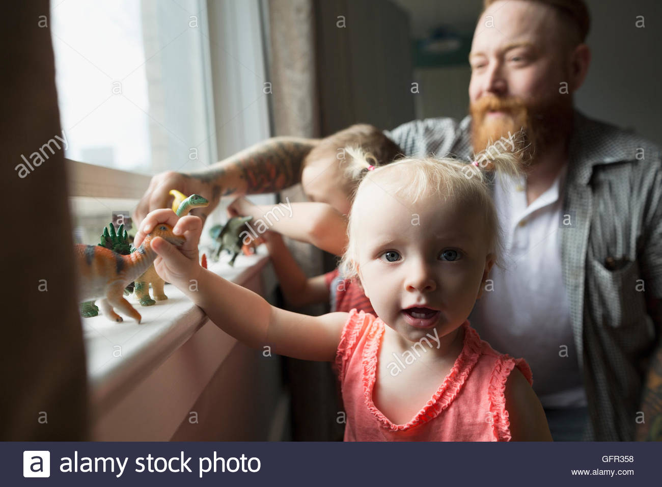 Portrait daughter and father playing with toy dinosaurs at windowsill Stock Photo