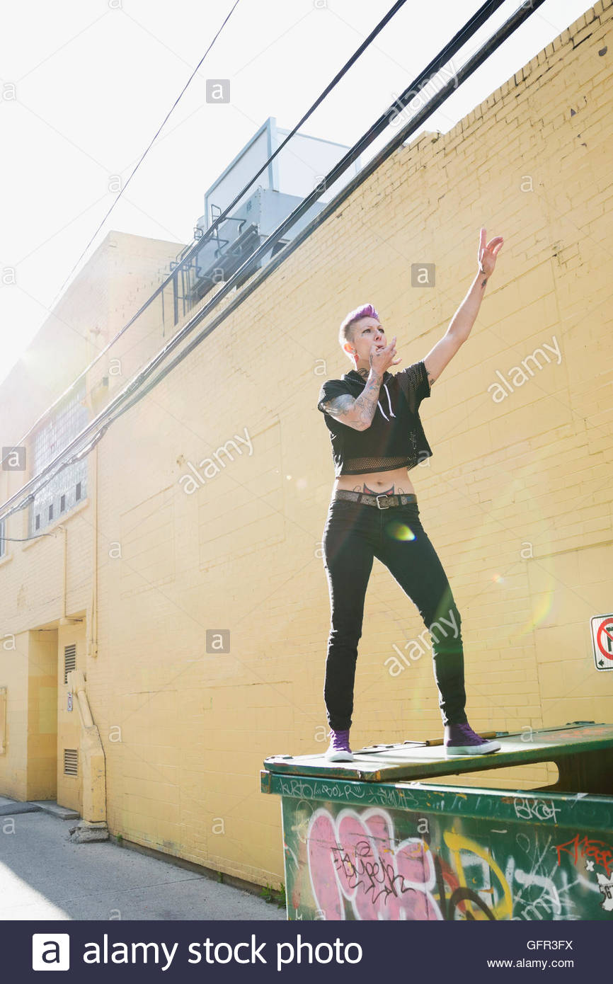 Cool young woman whistling on top of dumpster in sunny urban alley Stock Photo