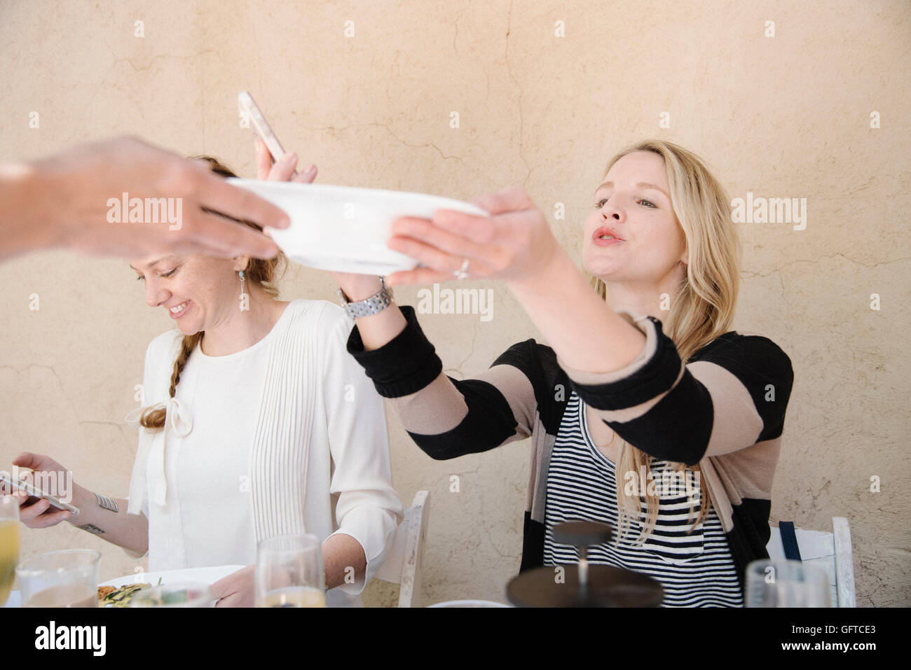Two smiling women sitting at a table one reaching for a plate of food Stock Photo